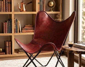 Könnte beinhalten: Ein dunkelroter Schmetterlingssessel aus Leder mit sichtbaren genähten Kanten und einem schwarzen Metallrahmen. Der Sessel hat eine geschwungene Sitzfläche im Sling-Stil. Das Wort PORRYS ist in Gold auf dem oberen rechten Teil der Rückenlehne gedruckt.