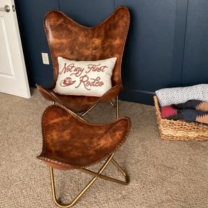 May include: A brown leather butterfly chair and matching ottoman with gold metal frames. A rectangular white throw pillow rests on the chair, featuring red embroidered text that reads Not my First Rodeo and a small red cowboy hat graphic.