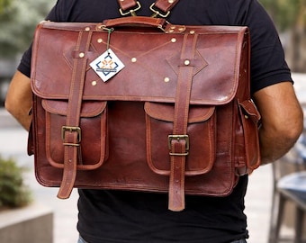 May include: A brown leather backpack with a large flap, two front pockets with buckle closures, and brass hardware. The bag features adjustable shoulder straps, side pockets, and a small white tag with a blue and orange logo attached to the top.