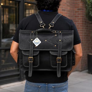 Può includere: Uno zaino in pelle nera con cuciture bianche a contrasto, dotato di due tasche frontali con fibbie in ottone, una maniglia superiore e spallacci regolabili. Un'etichetta bianca a forma di rombo con testo blu è fissata alla patta anteriore.
