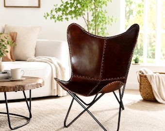 May include: A dark brown leather butterfly chair featuring a deep, curved seat with visible stitched seams and a black metal frame. The chair has a high backrest and a sling-style design supported by a crossed base, providing a structured seating form.