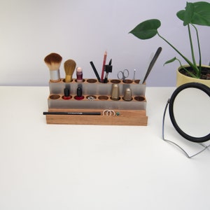 Puede incluir: Un organizador de madera con un inserto de metal plateado, que contiene varios pinceles de maquillaje, botellas de esmalte de u&ntilde;as y otros art&iacute;culos de belleza. El organizador tiene dos niveles y est&aacute; sobre una superficie blanca.