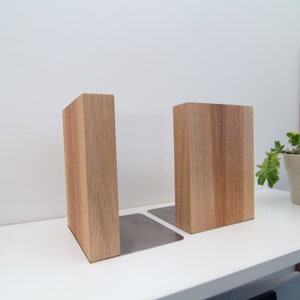 May include: Two wooden bookends with a natural wood grain finish. Each bookend has a rectangular wooden block attached to a silver metal base. The bookends are designed to hold books upright on a shelf or desk.