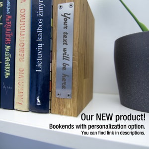 May include: Wooden bookends with a metal plate that reads "Your text will be here." The bookends are holding up several books with colorful spines. A potted plant is visible in the background.