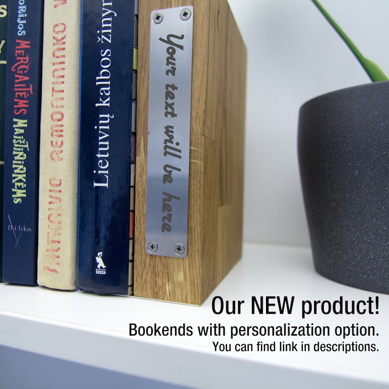 May include: Wooden bookends with a metal plate that reads "Your text will be here." The bookends are holding up several books with colorful spines. A potted plant is visible in the background.