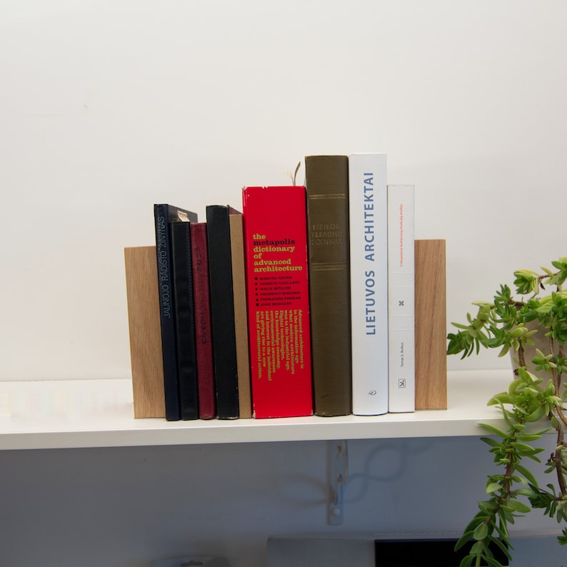 May include: A collection of books of varying sizes and colors, including red, black, and white spines, are held between two wooden bookends. The books are arranged on a white shelf, with a small green plant visible on the right.