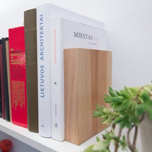 May include: A wooden bookend in a light oak finish stands next to a row of books on a white shelf. The books have various colors and titles, including "LIETUVOS ARCHITEKTAI" and "MIESTAS". A green plant is in the foreground.