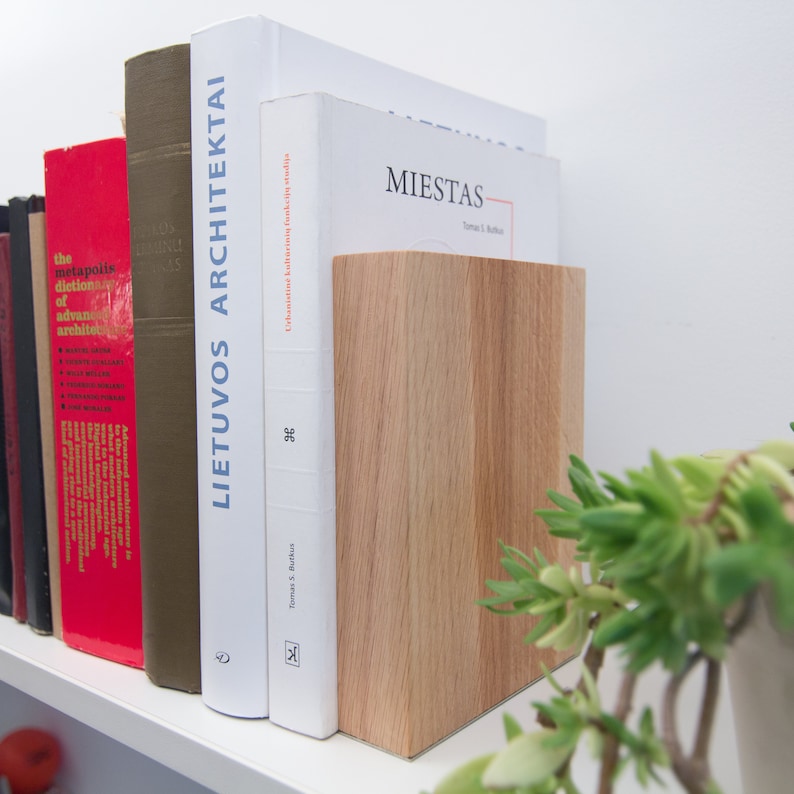 May include: A wooden bookend in a light oak finish stands next to a row of books on a white shelf. The books have various colors and titles, including "LIETUVOS ARCHITEKTAI" and "MIESTAS". A green plant is in the foreground.