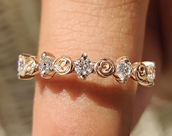 May include: A gold ring with a floral design, featuring small diamonds set in a repeating pattern. The ring is shown on a finger, with a neutral-toned nail polish.