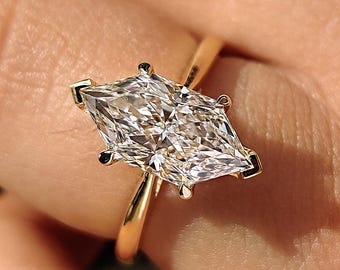 May include: A marquise-cut diamond engagement ring with a gold band. The diamond is set with six prongs, and the band is thin and polished. The ring is shown on a person's finger.