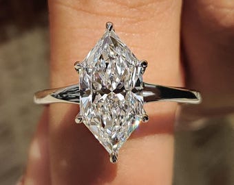 May include: A marquise-cut diamond engagement ring with a thin, silver band. The diamond is clear and faceted, reflecting light. The ring is held up by a person's finger, showing the ring's design and sparkle. The ring is a symbol of commitment and love.