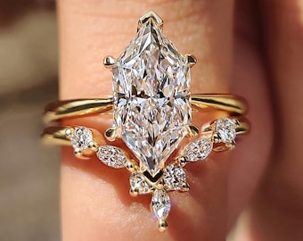 May include: A gold engagement ring set with a large, clear, diamond-shaped central stone. The ring is paired with a delicate band and a curved, diamond-studded band. The setting has a warm gold colour, creating a layered effect.