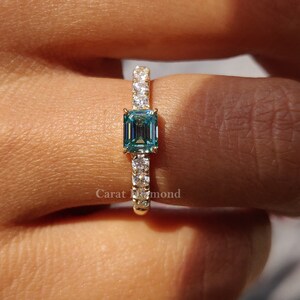 May include: A gold ring featuring a blue emerald-cut gemstone surrounded by smaller diamonds. The ring is worn on a finger.