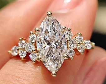 May include: A marquise-cut diamond engagement ring with a gold band. The ring features a large, central diamond and smaller diamonds clustered around it. The ring is held up by a person's hand.