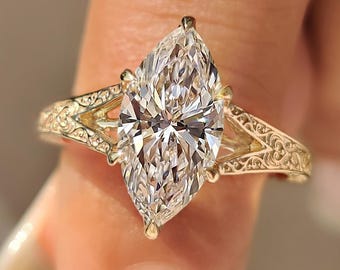 May include: A marquise-cut diamond ring featuring a clear, faceted center stone held by prongs. The gold band displays an intricate, engraved scrollwork pattern along the shoulders, creating a detailed texture on the metal surface.