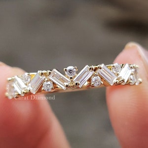 Puede incluir: Un anillo de banda de oro con una fila de diamantes talla baguette dispuestos en forma de chevron. El anillo está sobre un fondo blanco.
