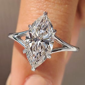 Puede incluir: Un anillo de compromiso con diamante talla marquesa y diseño de caña dividida. El diamante está engastado con garras y tiene muchas facetas. El anillo es de metal blanco, probablemente platino u oro blanco.