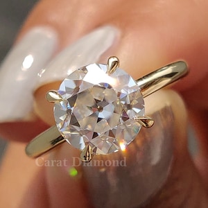 May include: A gold engagement ring with a large, round, clear diamond. The ring is on a finger and the diamond is sparkling.