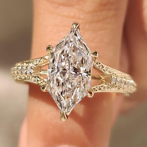 Puede incluir: Un anillo de oro con una gran piedra central de talla marquesa sujeta por seis garras. La banda muestra intrincados grabados de filigrana en los laterales y está acentuada con pequeñas piedras redondas incrustadas en los hombros.