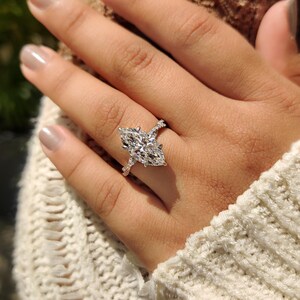 May include: A marquise-cut diamond engagement ring. The ring features a large, elongated diamond as the centerpiece, set on a band with smaller accent diamonds. The ring is shown on a hand wearing a white sweater.