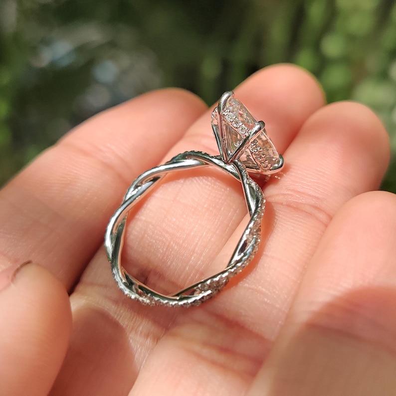 May include: A silver engagement ring with a large, clear, pear-shaped stone. The band has a twisted design, set with smaller stones. The ring is held in a hand, with a blurred green background.
