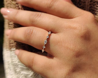 May include: A delicate gold ring adorned with a series of sparkling diamonds. The ring features a mix of round and marquise-cut stones, set in a linear pattern along the band. The ring is shown on a finger, showcasing its elegant design and subtle sparkle.