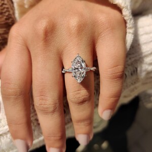 May include: A marquise-cut diamond ring. The ring showcases a large, clear diamond set in a delicate, twisted band. The band is embellished with smaller accent diamonds, adding sparkle. The ring is shown on a hand with painted nails.