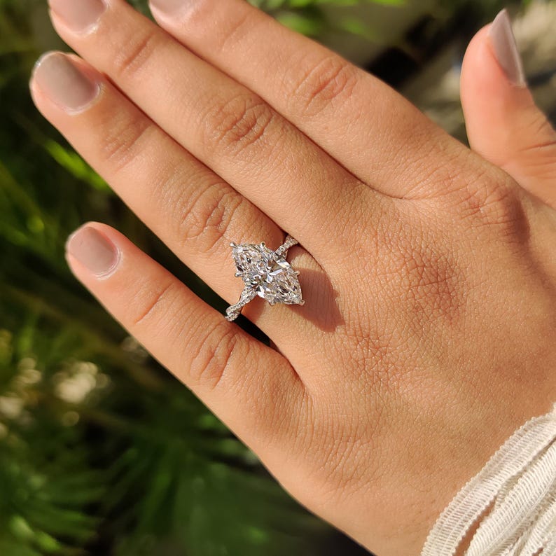 May include: A marquise-cut diamond engagement ring with a twisted band. The large, clear diamond is the focal point, set in a prong setting. The band is adorned with smaller diamonds. The ring is on a hand with neutral nail polish.