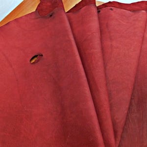 Puede incluir: Primer plano de varias hojas de cuero rojo oscuro, abanicadas para mostrar su textura y color. Una hoja tiene un agujero ovalado. El cuero parece suave y flexible, con una superficie ligeramente irregular.