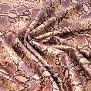 May include: Close-up of a fabric with a snakeskin pattern. The fabric has a mix of brown, tan, and purple tones, with a detailed print that mimics the scales of a snake. The fabric is draped, showing its texture and flexibility.
