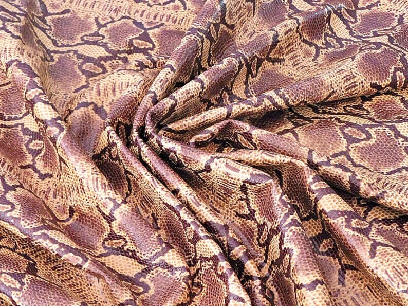 May include: Close-up of a fabric with a snakeskin pattern. The fabric has a mix of brown, tan, and purple tones, with a detailed print that mimics the scales of a snake. The fabric is draped, showing its texture and flexibility.