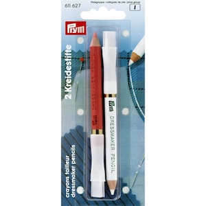 May include: Two dressmaker pencils, one red and one white, with a brush on the end of each. The pencils are packaged together in a clear plastic wrapper with the brand name "Prym" and the text "Dressmaker Pencil" printed on the packaging.