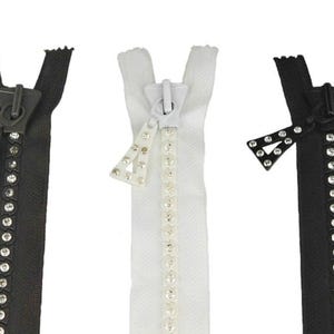 Può includere: Tre cerniere decorative in bianco e nero. Ogni cerniera presenta un tirazip unico ornato di strass e una fila di strass lungo i denti. Le cerniere sono esposte su uno sfondo bianco.