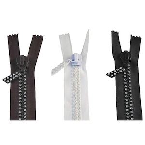Puede incluir: Tres cremalleras en marrón, blanco y negro. Cada cremallera tiene una lengüeta a juego con pedrería decorativa. Los dientes de la cremallera también están coordinados por color. Las cremalleras se muestran sobre un fondo blanco.