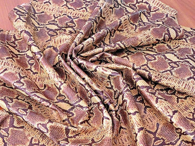 May include: A close-up of a fabric with a snakeskin pattern. The fabric has a mix of brown, tan, and black colours. The material is draped, showing the texture and pattern of the snakeskin design. The fabric is likely used for clothing or accessories.