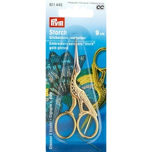 May include: Gold-plated stork-shaped embroidery scissors measuring 9 cm in length. The scissors are packaged in a clear plastic blister pack on a blue cardboard backing featuring the Prym logo and the text Storch, Embroidery scissors Stork.