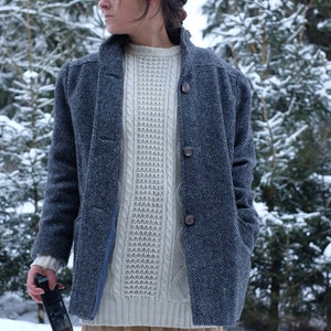 May include: A woman wearing a blue herringbone coat over a white cable knit sweater and a beige skirt. She is holding a black smartphone in her left hand.