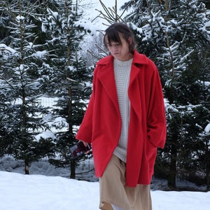 May include: A person wearing a bright red coat, a white sweater, and a tan skirt, walking in a snowy forest. The coat has a wide collar and is open in the front. The person is holding a phone.