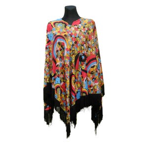 May include: A colorful, patterned poncho with a V-neck and black fringe. The fabric features a vibrant mix of red, yellow, blue, and green floral and abstract designs. The poncho is displayed on a black mannequin head.