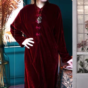 May include: A long, burgundy velvet dress with a mandarin collar and long sleeves. The dress features decorative frog closures down the front and a pendant necklace. The dress is a rich, deep red color, and the fabric has a soft, luxurious sheen.