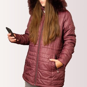 Puede incluir: Chaqueta acolchada burdeos con capucha ribeteada de piel sintética. La chaqueta tiene cremallera completa, dos bolsillos laterales y un pequeño dije en la cremallera. La persona sostiene un teléfono inteligente negro.
