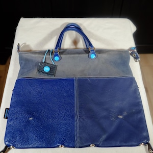 Op de afbeelding: Een tweekleurige leren tote bag in blauw- en grijstinten. De tas heeft een gestructureerd ontwerp met een handvat aan de bovenkant, een klein label en zilverkleurige hardware. De tas is verdeeld in twee secties, met een donkerder blauw aan de onderkant en een lichter grijs aan de bovenkant.