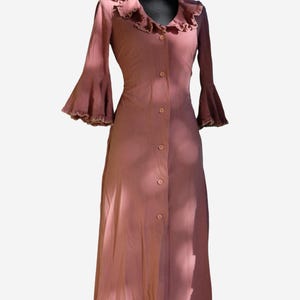 Könnte beinhalten: Ein staubrosa Kleid mit Knopfleiste und Rüschenkragen. Das Kleid hat lange Ärmel mit ausgestellten Manschetten. Das Kleid wird auf einer schwarzen Schaufensterpuppe präsentiert.