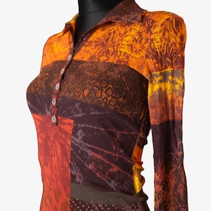 Può includere: Una camicia a maniche lunghe con colletto e un vivace design patchwork. La camicia presenta un'abbottonatura frontale e un mix di colori caldi, tra cui arancione, marrone e rosso. Il tessuto ha un aspetto strutturato, aggiungendo interesse visivo all'indumento.