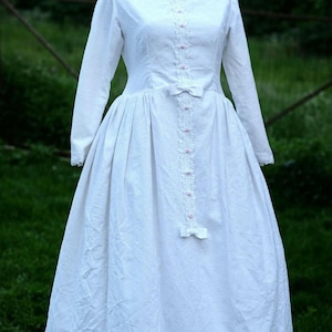 May include: A white dress with a long sleeve and a pink lace trim down the front. The dress has a full skirt and a small bow at the waist.