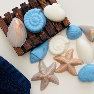 May include: A collection of seashell-shaped soaps in various colors, including blue, white, and tan, arranged on a wooden soap dish. Some shells have metallic gold accents. A blue towel is in the lower left corner.