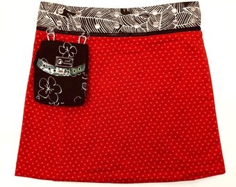Puede incluir: Una falda roja con un patrón de estrellas blancas y un bolsillo negro con un diseño floral. La cintura tiene una banda estampada en blanco y negro con ojales plateados.