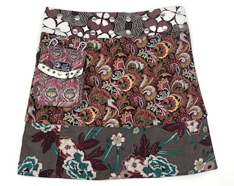 May include: A wrap skirt with a patchwork design. The skirt features a pocket with a peacock feather pattern, a paisley print, and a floral print. The top band has a heart and dot pattern. The skirt has silver grommets.