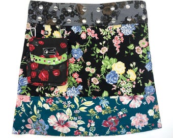 May include: A multi-colored patchwork skirt with a floral print. The skirt has a black fabric with a floral print, a teal fabric with a floral print, and a gray fabric with polka dots. The skirt has a small pocket with a green and red floral print and the text "Love Revolution" on it.