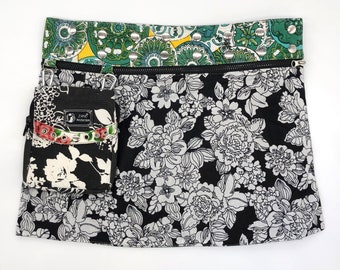 Puede incluir: Una falda negra con estampado floral blanco y una cintura estampada en verde, amarillo y azul con tachuelas plateadas. Un pequeño bolso negro con cremallera y estampado floral blanco está unido a la falda.